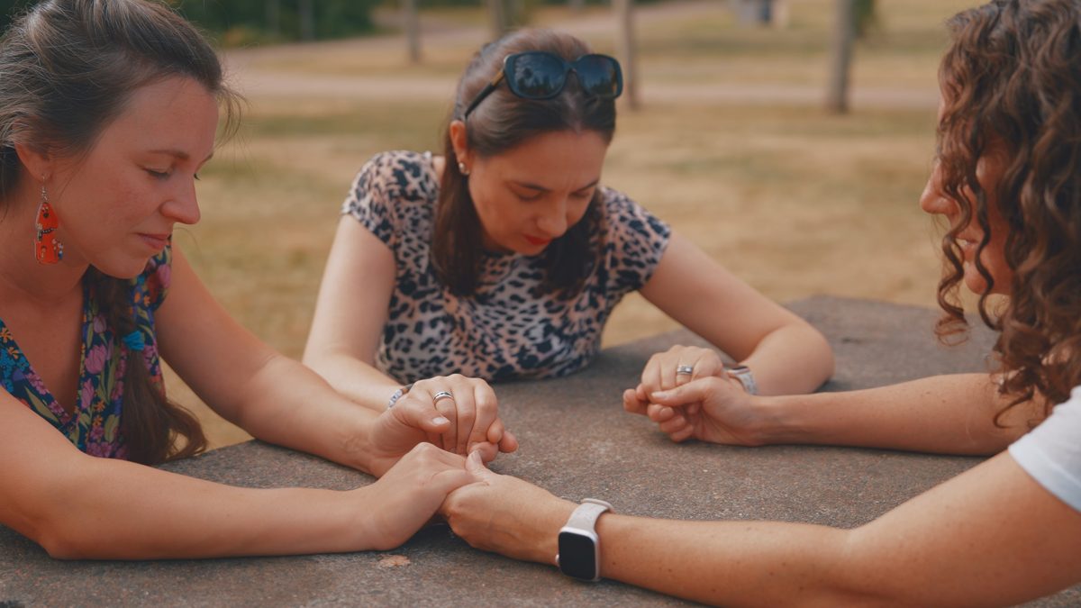 Three moms holding hands and praying together at a park picnic table