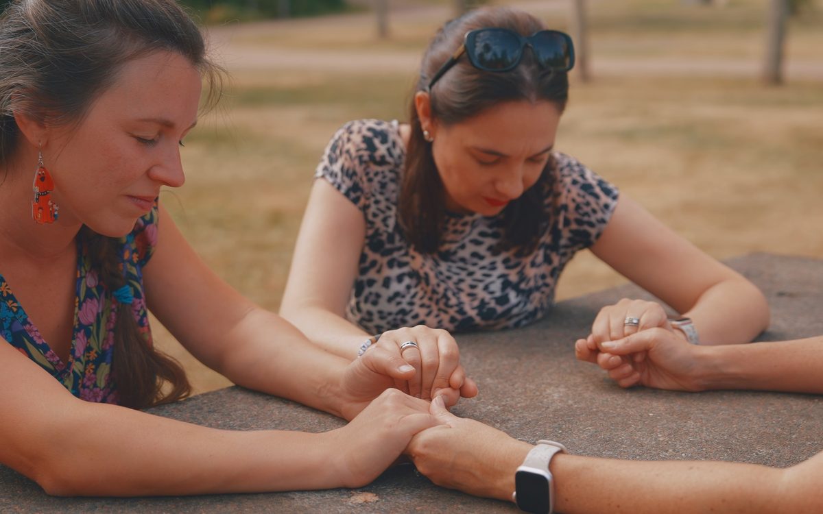 Three moms holding hands and praying together at a park picnic table